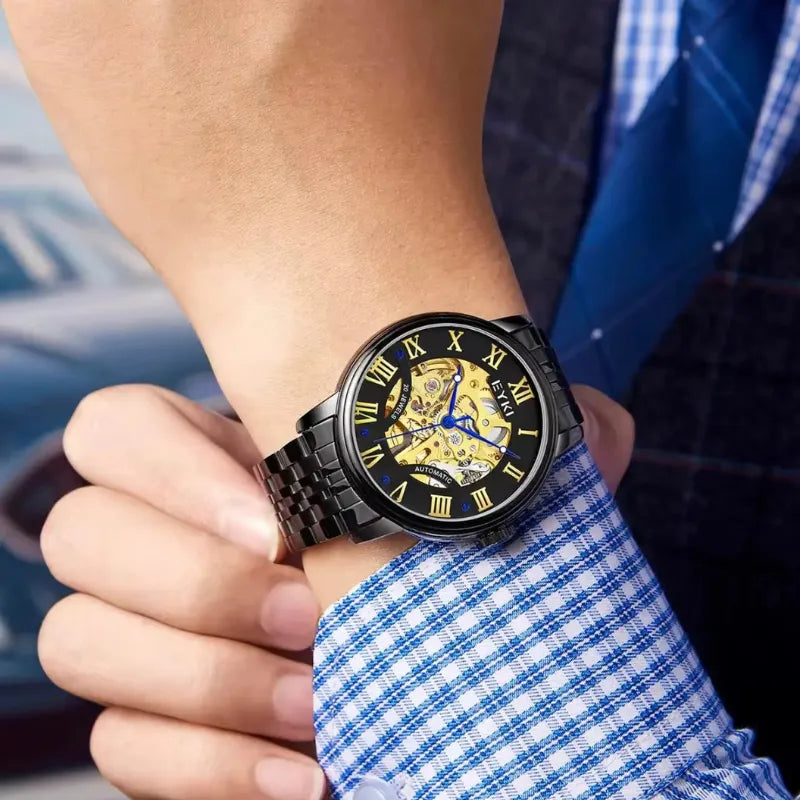 Person wearing a black wristwatch with a detailed face, blurred car and blue checkered shirt in the background