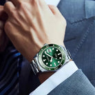 Green watch with silver case on a person's wrist wearing a suit.