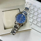 Silver watch with blue face on a white surface next to a laptop