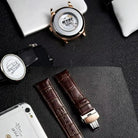 Watch with open face, brown leather strap, and smartphone on a dark textured surface